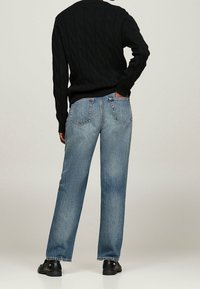 Dunkelblaue Jeans mit lässigem Schnitt, schmal zulaufenden Beinen und Gesäßtaschen. Getragen mit einem schwarzen, grobstrickigen Pullover und schwarzen Schuhen.