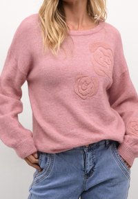 Rosa gestrickter Pullover mit langen Ärmeln, versehen mit strukturierten Rosenmotiven in einem helleren Ton. Kombiniert mit hellblauen Denim-Jeans.