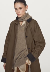 Brown outer coat with button closure, contrasting dark collar, over a beige knit sweater, lightweight beige shirt, and light brown pants.