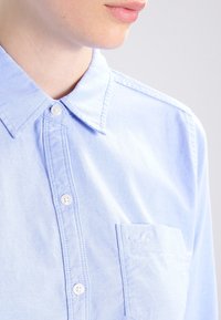 Camicia azzurra chiara con colletto standard, bottoni frontali e taschino sul petto con logo. Il tessuto appare morbido e liscio.