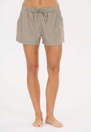 Jambes et pieds nus d'une personne portant un short beige clair à taille élastique avec un cordon à l'avant, debout sur un fond blanc.