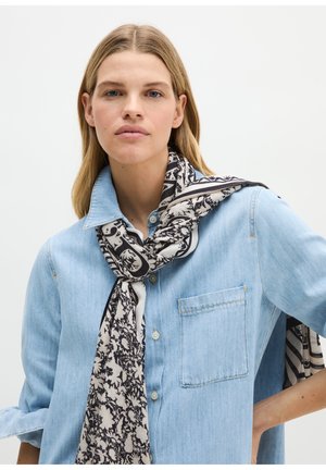 Donna con lunghi capelli biondi che indossa una camicia di jeans azzurro chiaro e una sciarpa floreale bianca e nera su uno sfondo uniforme.