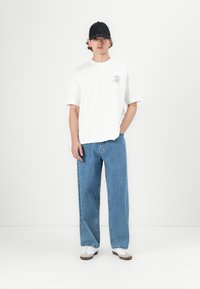 White casual t-shirt with short sleeves and a small graphic design on the left chest, paired with loose-fitting blue denim jeans and white shoes.