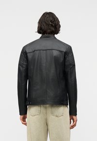 Person with shoulder-length dark hair wearing a black leather jacket and light beige pants, shown from the back against a white background.