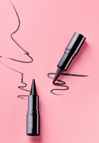 Deux crayons eye-liner noirs Lord & Berry sur fond rose avec des lignes noires tourbillonnantes et en zigzag dessinées.