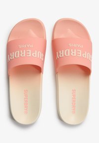 Un par de sandalias tipo slide de Superdry en tonos rosa y crema con suelas texturizadas y correas con la marca, mostradas desde arriba sobre un fondo blanco.
