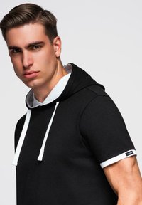 Mladý muž s krátkými hnědými vlasy, oblečený v černé mikině s krátkým rukávem, bílými šňůrkami a bílým lemem na rukávech, který se mírně dívá na stranu.