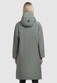 Cămașă lungă, gri, impermeabilă, cu glugă; dispune de o talie strânsă, fante laterale și un logo pe spate. Materialul are o textură netedă.