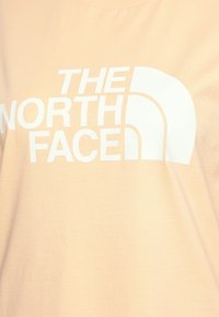 Světle broskvová bavlněná trička s velkým bílým logem "THE NORTH FACE" a půlkruhovým emblémem na horní levé straně.