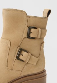Botte en daim beige à la cheville avec des accents de double boucle, un orteil arrondi et une semelle en bois. Détails de couture le long des coutures.