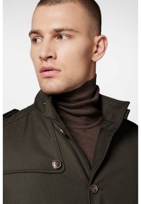 Khaki trencz z wysokim kołnierzem i mosiężnymi guzikami, nałożony na brązowy sweter z prążkowaną fakturą. Gładka tekstura materiału.