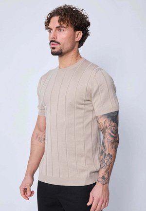 Homme aux cheveux bouclés et à la barbe portant une chemise tricotée beige à manches courtes, un pantalon noir, avec des tatouages floraux et de silhouettes stylisées sur les bras et le cou.