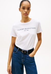 Balta medvilnės marškinėliai su "TOMMY HILFIGER EST. 1985" jūros mėlyna spausdinimu, derinami su aukštu juosmeniu melsvomis džinsinėmis kelnėmis ir juodu diržu.