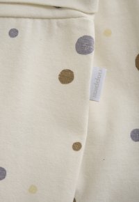 Cremfarbener Stoff mit großen braunen, lila und gelben Punkten sowie einem weißen Stoffetikett mit prägierter Schrift an der Naht.