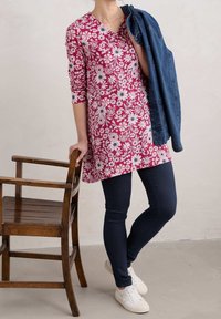 Czerwony kwiatowy tunik z rękawami 3/4, dekoltem w serek, zestawiony z ciemnymi dżinsami i białymi trampkami, trzymając dżinsową kurtkę. Obok drewniane krzesło.