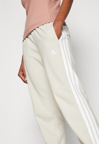 Beige joggebukser har hvite side-striper og et brodert logo. Laget av myk tekstil med elastisk midjebånd og lommer.