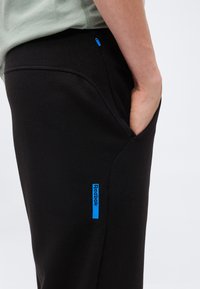 Άτομο που φοράει μαύρο παντελόνι Reebok με το χέρι στην πλαϊνή τσέπη, που διαθέτει ένα μικρό γαλάζιο φερμουάρ και μπλε λωρίδα με το λογότυπο Reebok.