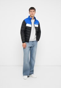 Polstret jakke med en blå og hvid øverste del, sort nederste del, høj krave og båret over en grå sweater med lyseblå jeans.