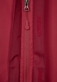 Röd tyg med dragkedja; har kardborreöverdrag bredvid dragkedjan. Slät textur, minimala designelement, praktisk för utomhusbruk.