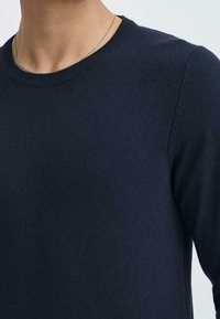 Dunkelblauer, gestrickter Pullover mit klassischem Rundhalsausschnitt und Rippdetails an den Ärmeln und am Saum. Weiche Textur mit taillierter Silhouette.