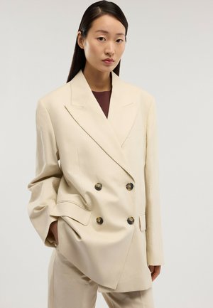 Mujer con cabello largo y oscuro que lleva una americana oversize color crema con doble botonadura y pantalones a juego, de pie frente a un fondo claro y sencillo.