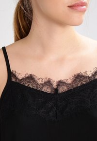 Kvinnas hals och övre bröst bär en svart linne med delikat blommig spetskant och tunna axelband.