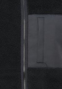 Black fleece jacket featuring a smooth fabric section, central zipper with metal teeth, and a patch pocket on the left side.