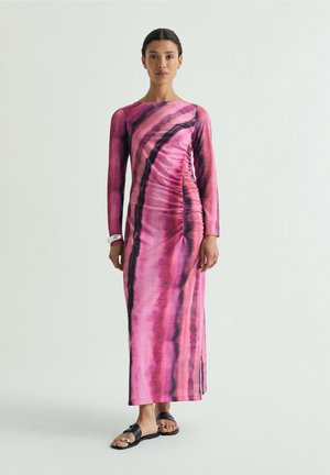 Mujer de pie con un vestido largo de manga larga en color rosa y negro con efecto tie-dye, fruncido lateral y sandalias planas negras sobre un fondo liso.