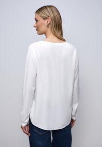 Blusa bianca a maniche lunghe con scollatura arrotondata, texture liscia, orlo leggermente curvato e dettagli con bottoni ai polsini.