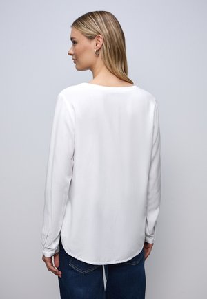 Blouse blanche à manches longues avec un col rond, texture lisse, ourlet légèrement incurvé et accents de boutons aux poignets.