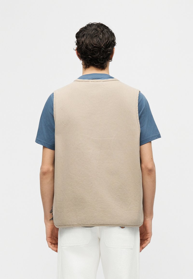 Hombre con cabello rizado y oscuro que lleva un chaleco sin mangas beige sobre una camisa azul de manga corta y pantalones blancos, de espaldas sobre un fondo liso.