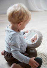 Peuter in grijs shirt zittend op de vloer, houdend een zacht speelgoed vast met een ronde spiegel die hun gezicht weerspiegelt.