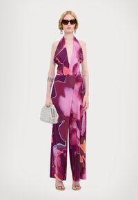 Tuta senza maniche con scollatura a halter in un motivo floreale viola e rosa, con gambe ampie. La modella tiene una borsa a tracolla di metallo argentato, indossando occhiali da sole marroni.