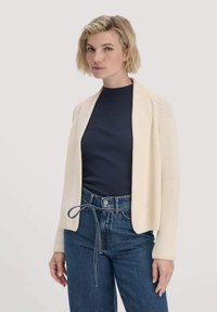 Strickcreme-Cardigan mit offenem Vorderteil, strukturiertem Finish, langen Ärmeln, getragen über einem marineblauen Top und hoch taillierten blauen Jeans.