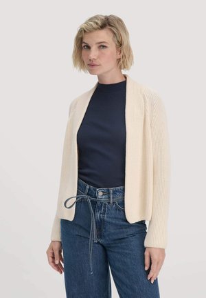 Gebreide crèmekleurige cardigan met open voorkant, textuurafwerking, lange mouwen, gedragen over een marineblauw topje en hoogdelige blauwe jeans.