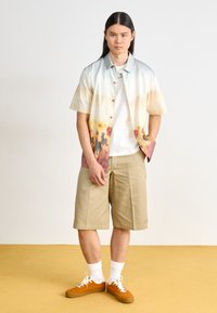 Short-sleeve shirt in gradient colors with a beach scene, paired with beige shorts and orange sneakers, white socks. Room with yellow floor.