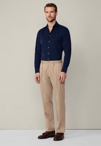 Camisa de manga larga azul marino con cierre de botones, combinada con pantalones de pliegues en un beige claro. Unos zapatos marrones completan el conjunto. Fondo neutro.