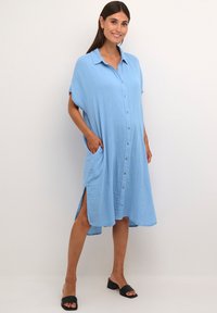 Mėlyna marškininė suknelė iš lengvo audinio, su trumpomis rankovėmis, šoninėmis kišenėmis ir laisva siluetu, su skeltukais apačioje.