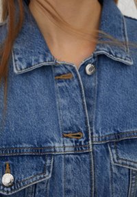 Section d'une veste en denim bleu avec col, bouton et coutures visibles, portée par une personne à la peau claire et aux mèches de cheveux bruns.