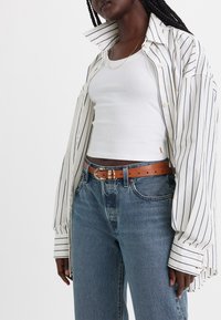 Lang gestreepte shirt in wit en marineblauw, witte tanktop, bruine riem met een gouden gesp, en een spijkerbroek in een ontspannen fit.