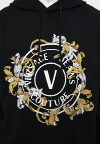 Juoda liemenė su apvaliu grafiniu dizainu baltos ir aukso spalvų, kuriame užrašyta "VERSACE JEANS COUTURE" ir puošni lapų motyvas supantis didelį "V".