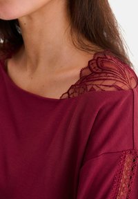 Top bordeaux avec des accents de dentelle semi-transparente aux épaules et aux manches, texture lisse, coupe décontractée et bordure en dentelle décorative.
