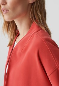 Veste rouge légère avec une texture lisse et un col doux, présentant une coupe décontractée et des détails de couture minimaux.