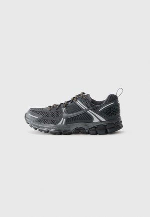 Zapatilla de running para hombre en malla negra con acentos grises. Presenta una suela texturizada, punta redondeada y elementos reflectantes. Diseño con cordones y tirador en el talón.