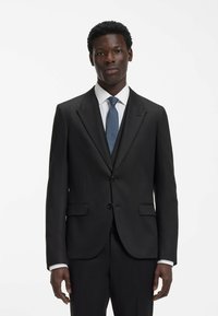 Abito nero con rever a incavo e due bottoni, completato da una camicia bianca e una cravatta blu strutturata. Tessuto liscio e vestibilità sartoriale.