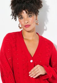 Roter Strickcardigan mit V-Ausschnitt, der mit einer Knopfleiste, offenem Muster und langenärmeln gestaltet ist, präsentiert vor einem neutralen Hintergrund.