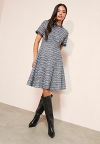 Rövid ujjú, testhez álló és szoknyarészben bővülő ruha texturált tengerészkék és fehér anyagból; rojtos szegélyekkel és kerek nyakkivágással. Magas fekete csizmával viselve.