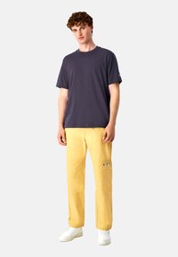 Mørk grå t-shirt til mænd med rund hals, parret med lysegule, lige bukser med et lille logo. Hvide sneakers fuldender looket.