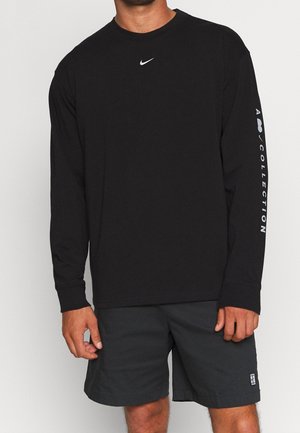 Homme portant un t-shirt noir à manches longues Nike avec "A Collection" sur la manche et un short noir sur un fond uni.