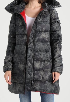 Frau trägt einen langen schwarzen Steppmantel mit einem dezenten Spitzenmuster, roter Innenfütterung und weitem Kragen, kombiniert mit blauen Jeans.